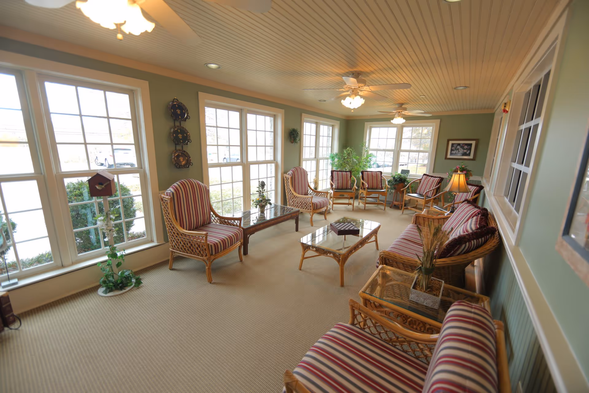 A bright and cozy sitting room with large windows letting in natural light. The room features wicker furniture with red and white striped cushions, including armchairs, a loveseat, and coffee tables with glass tops. Ceiling fans with lights hang from the beadboard ceiling, and there are plants and decorative items placed around the room.