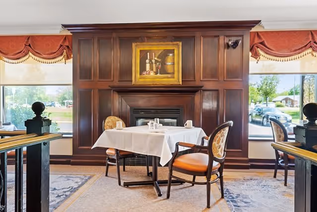 A cozy dining area with a table covered by a white tablecloth set for two, positioned in front of a dark wood-paneled wall with a fireplace. Above the fireplace hangs a painting of wine bottles and a barrel. The room has large windows with red valance curtains, allowing natural light to brighten the space.