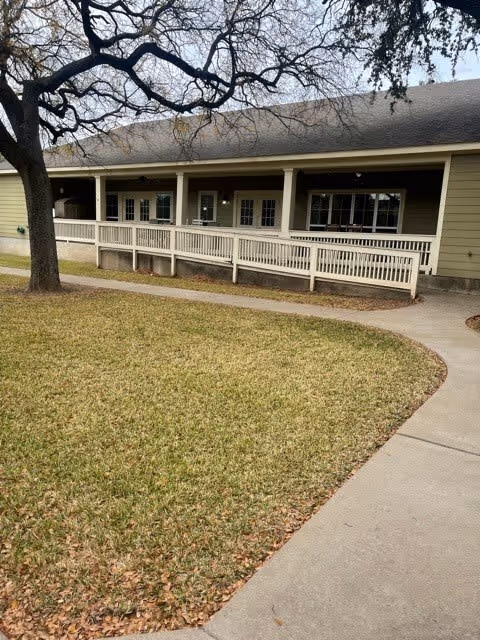 Exterior view of a single-story building with a covered porch and a wheelchair-accessible ramp. The building is surrounded by a lawn with a large tree and a curved concrete walkway leading to the entrance.