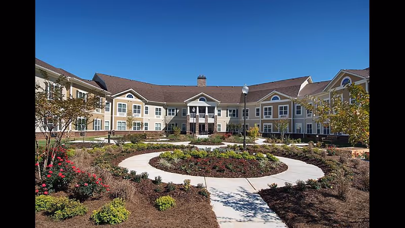 Exterior view of a senior living facility building with beige and white walls, multiple windows, and a brown roof under a clear blue sky. In front of the building is a landscaped garden with a circular walkway, various shrubs, and flowering plants.