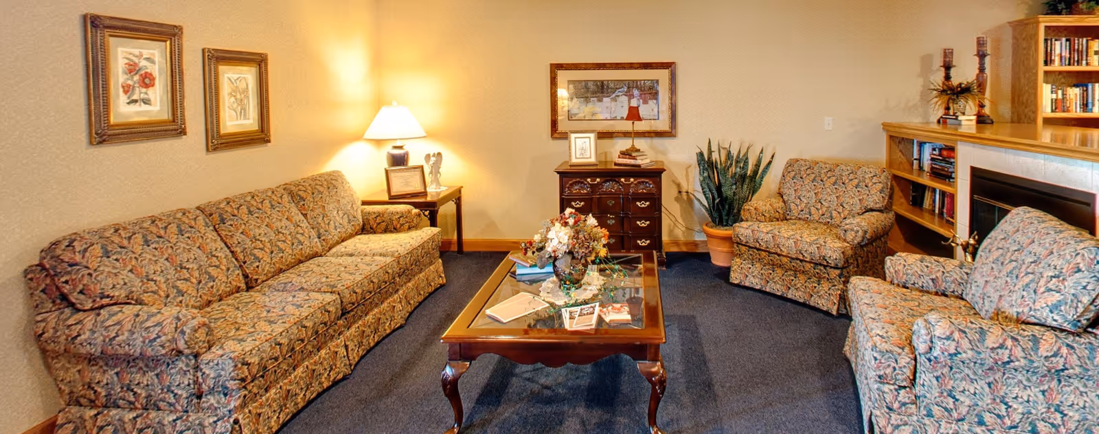 Cozy living room with patterned sofas and armchairs arranged around a wooden coffee table, lamp, artwork, and a bookcase.