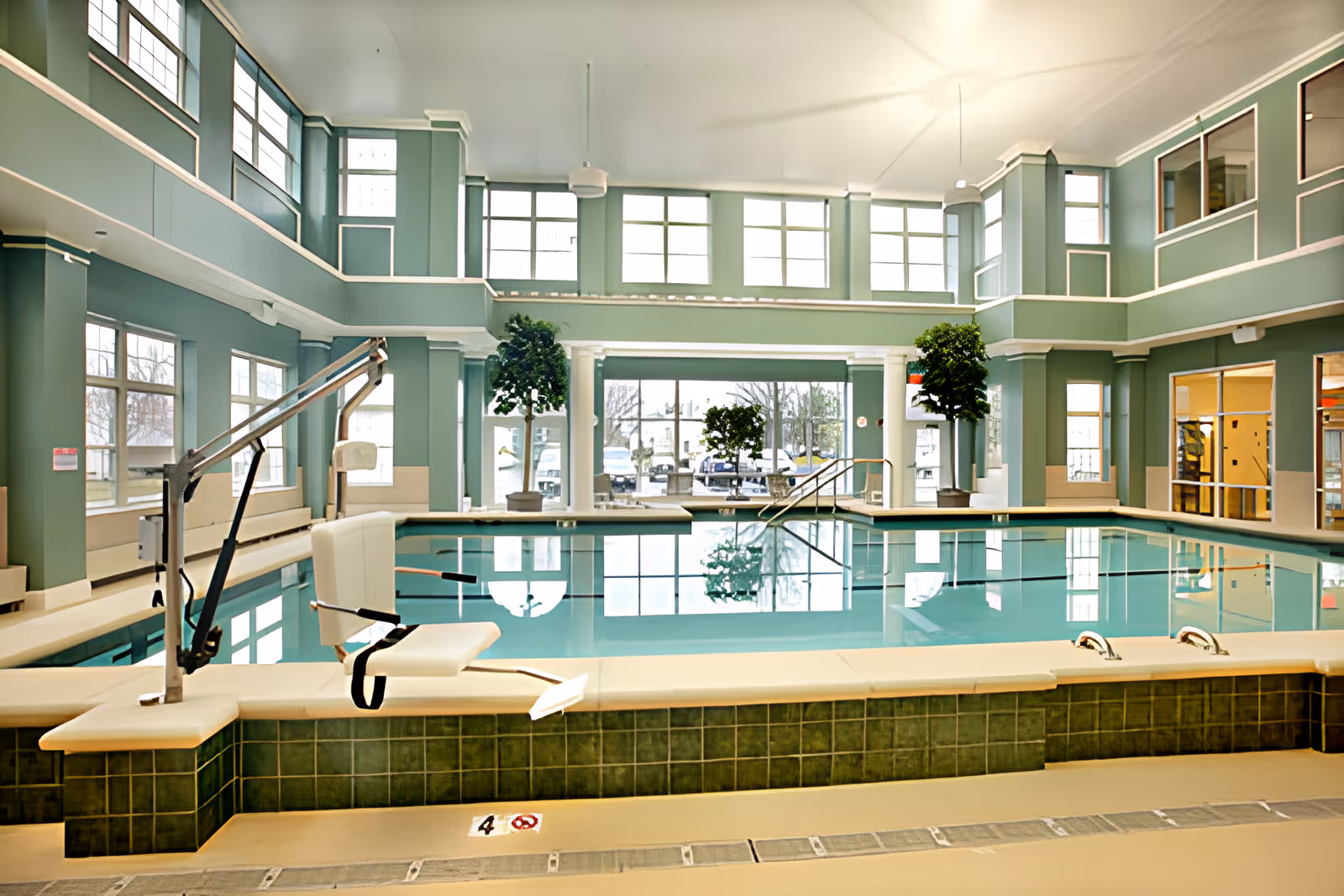 Indoor swimming pool area with large windows allowing natural light, green tiled pool edge, pool lift chair for accessibility, and potted plants near the windows.