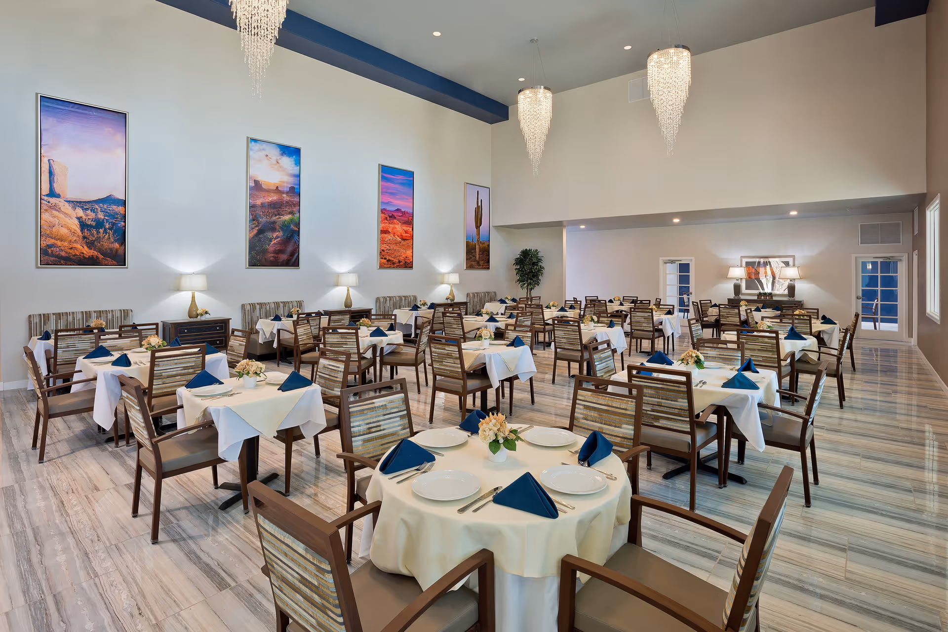 Spacious dining room with multiple tables set with white tablecloths and blue napkins, chandeliers, and landscape artwork on the walls.