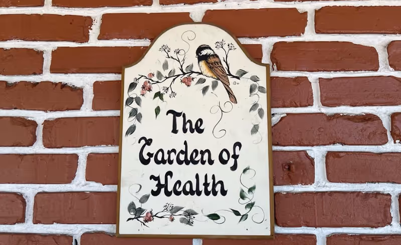 A decorative sign reading "The Garden of Health" with a painted bird mounted on a red brick wall.