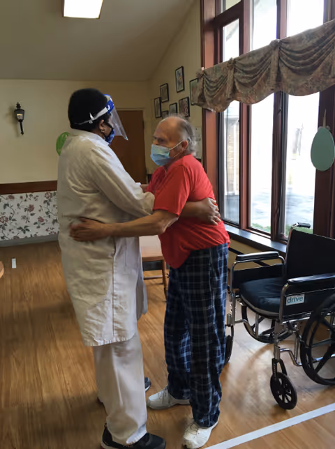 A healthcare worker wearing a white coat, face shield, and mask embraces an elderly man in a red shirt and plaid pants who is also wearing a mask. They are standing in a room with wooden floors, large windows with curtains, and a wheelchair nearby.