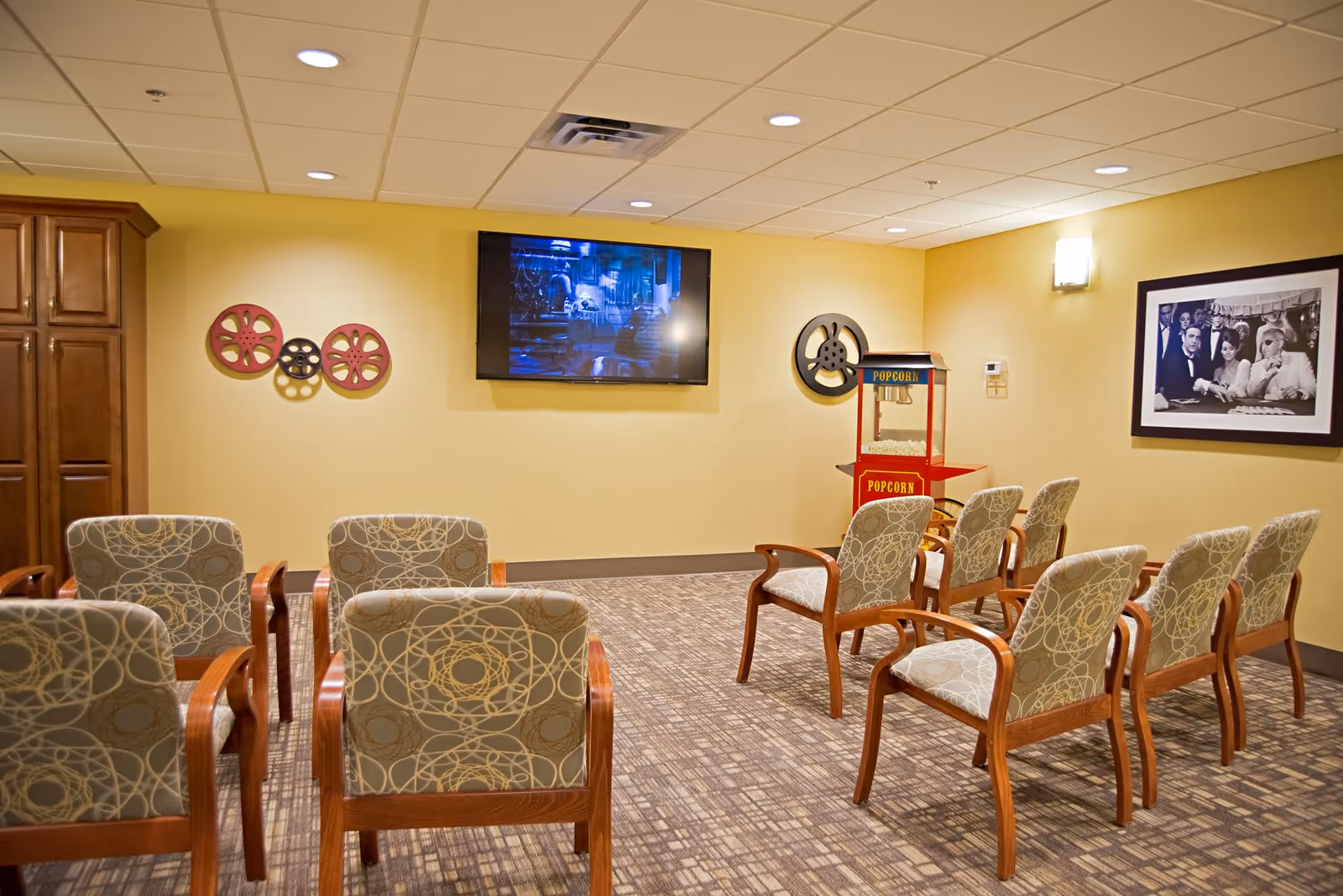 A small theater room with two rows of cushioned chairs facing a wall-mounted flat screen TV. The walls are painted yellow and decorated with film reel wall art and a black and white framed photo. A red popcorn machine is positioned in the corner of the room.
