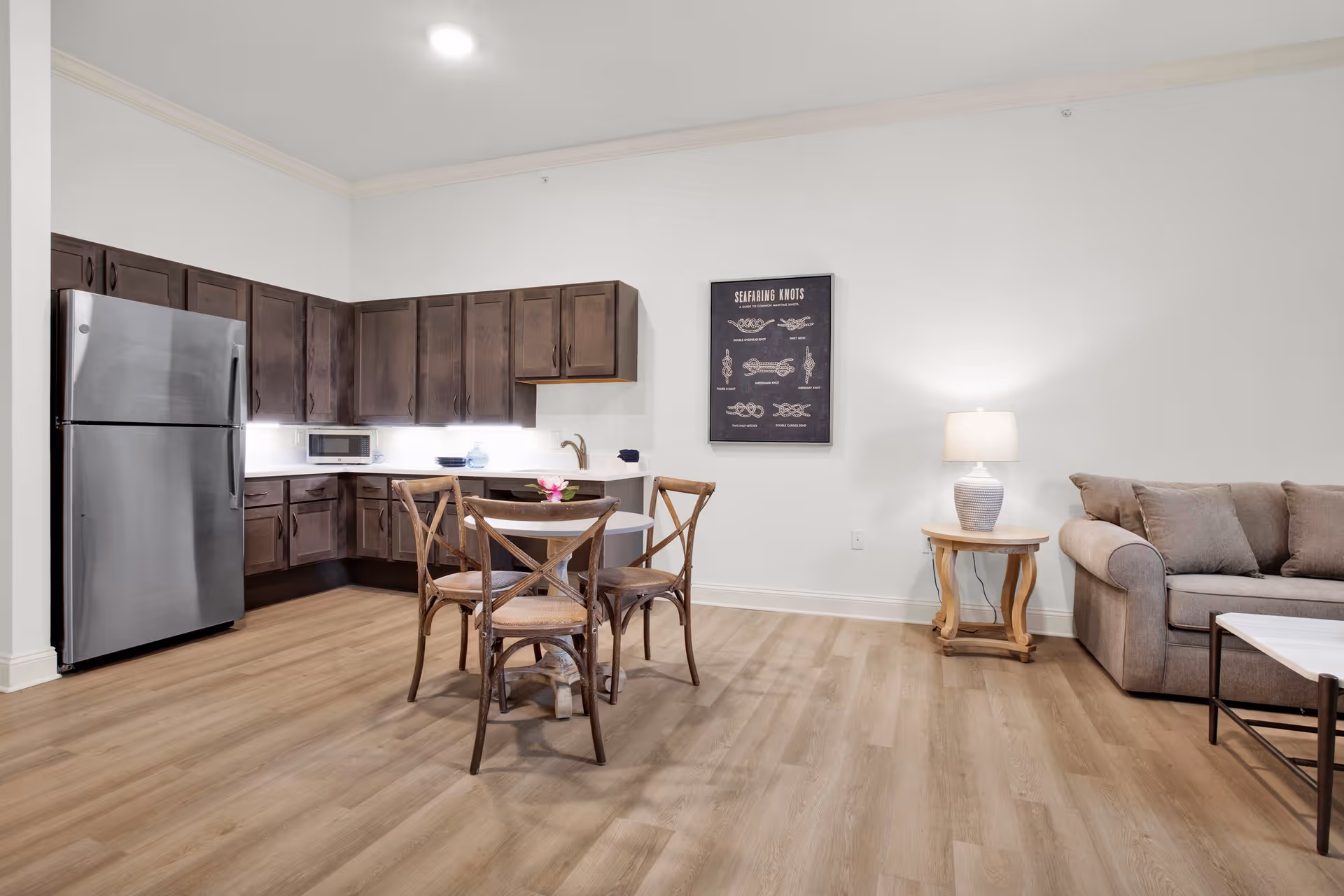 A modern open-concept living space featuring a kitchen with dark wooden cabinets, a stainless steel refrigerator, and a microwave. In front of the kitchen is a round dining table with four wooden chairs. To the right, there is a beige sofa with cushions, a wooden side table with a lamp, and a coffee table. A framed artwork titled 'Seafaring Knots' hangs on the wall above the side table. The room has light wood flooring and white walls.