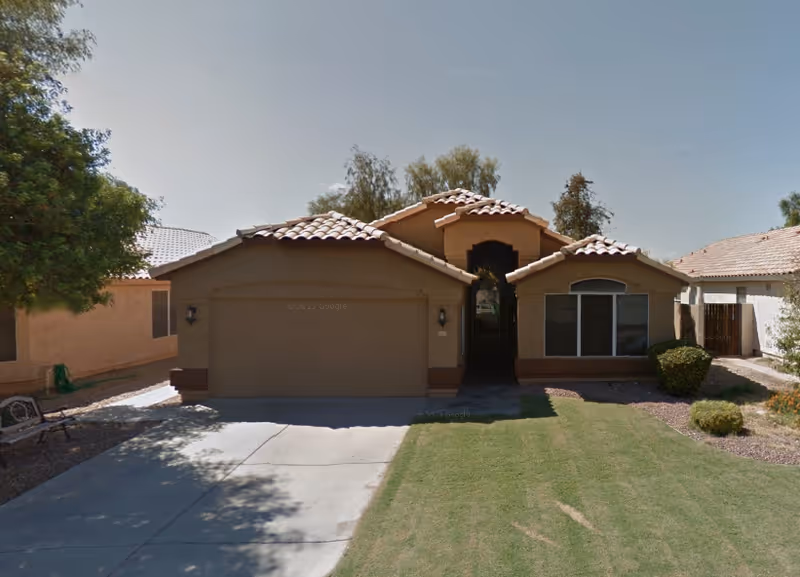 Front exterior view of a single-story house with a tiled roof, a two-car garage, a driveway, and a well-maintained lawn with some bushes and trees on either side.