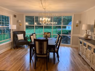 Dining room with a table and chairs under a chandelier and large windows overlooking a grassy yard.