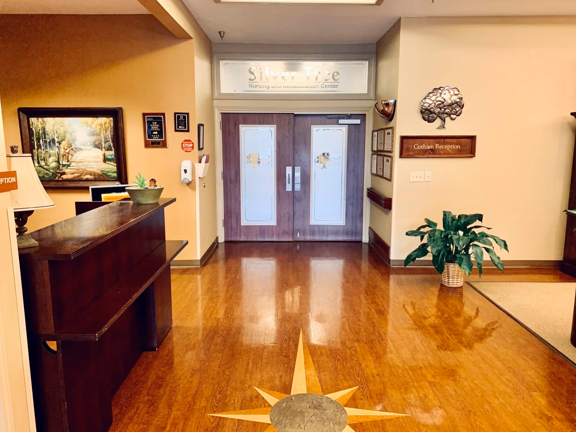 Reception area of Silver Tree Nursing and Rehabilitation Center featuring a wooden reception desk with a lamp and plant, polished wooden floor with a star design inlay, double wooden doors with frosted glass panels, a painting on the left wall, plaques and a hand sanitizer dispenser on the left wall, a plant in a basket on the right, and a sign reading 'Cotham Reception' with a decorative tree above it on the right wall.