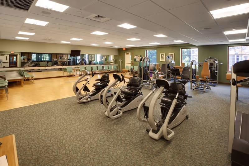A spacious fitness room with exercise equipment including three recumbent stationary bikes in the foreground and various weight machines in the background. The room has carpeted and wooden flooring sections, large mirrors on one wall, several windows letting in natural light, and green walls. Chairs are lined up along the mirrored wall.