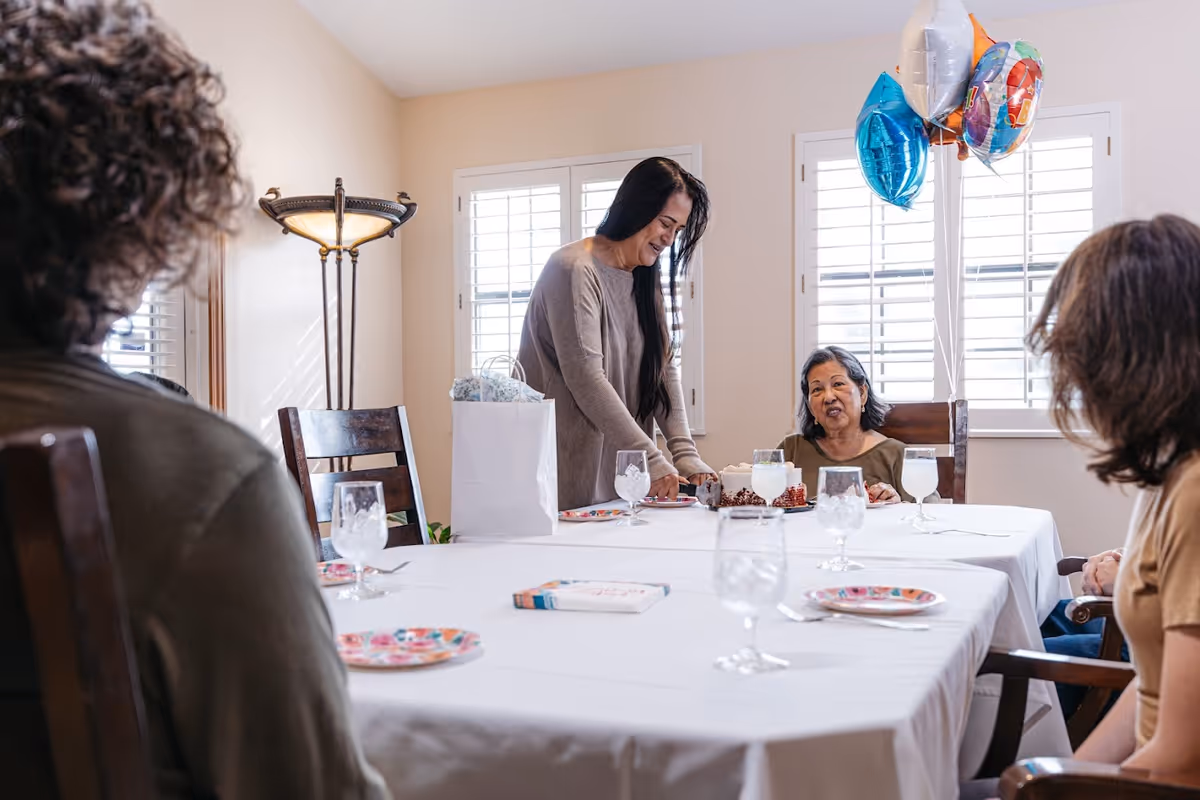 A group of people gathered around a dining table with a white tablecloth. One woman is standing and cutting a cake while others are seated, with balloons tied to a chair in the background near windows with white shutters.