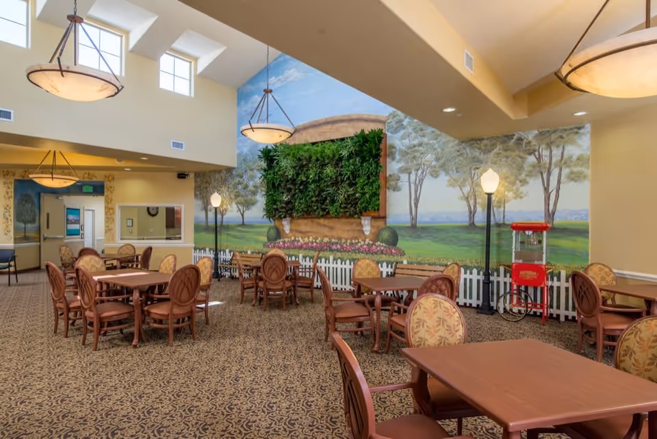 A spacious dining area with multiple wooden tables and chairs arranged on a patterned carpet. The walls feature a large mural depicting a garden scene with trees, a white picket fence, and a vertical garden with green plants. There are two tall lamp posts inside the room and a red popcorn machine near the wall. The ceiling has large hanging light fixtures and several windows near the top letting in natural light.
