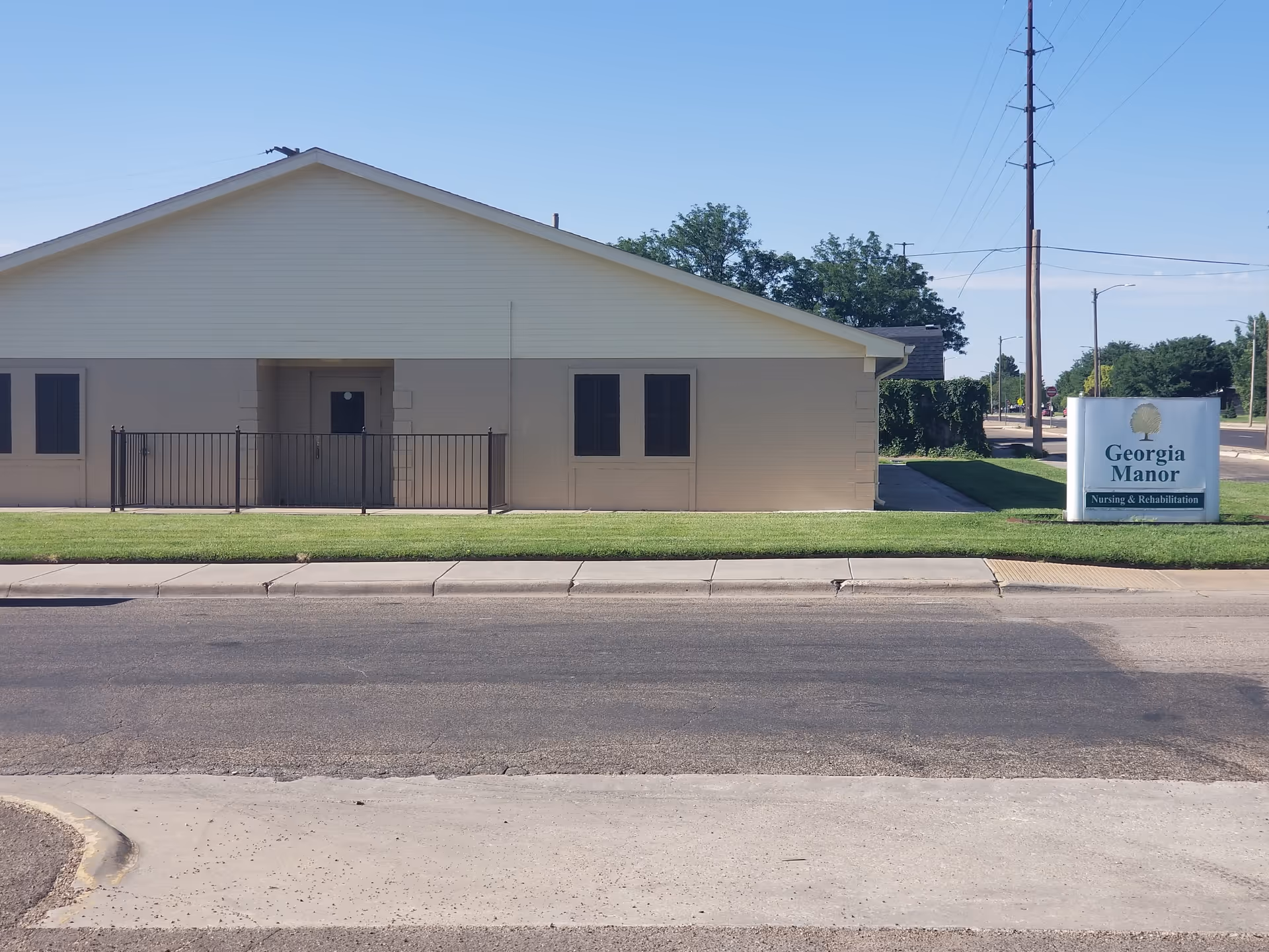 Exterior view of a single-story building with beige walls and a light-colored roof, identified as Georgia Manor Nursing & Rehabilitation by a sign on the right side near the street. The building is set behind a small grassy area and a sidewalk, with a clear blue sky overhead.