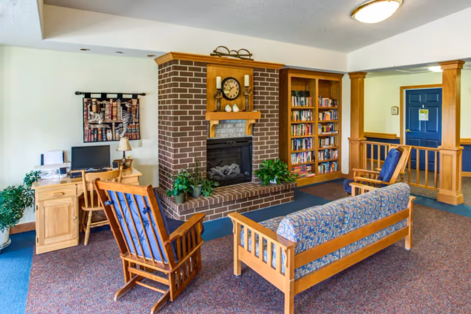 A cozy living room area with wooden furniture including a blue cushioned sofa and two matching chairs. There is a brick fireplace with a clock and decorative items on the mantel. A wooden bookshelf filled with books is next to the fireplace. A small wooden desk with a computer and lamp is positioned against the wall, and there are several potted plants around the room. The walls are light-colored, and the carpet is a mix of blue and red tones. A blue door and wooden railing are visible in the background.