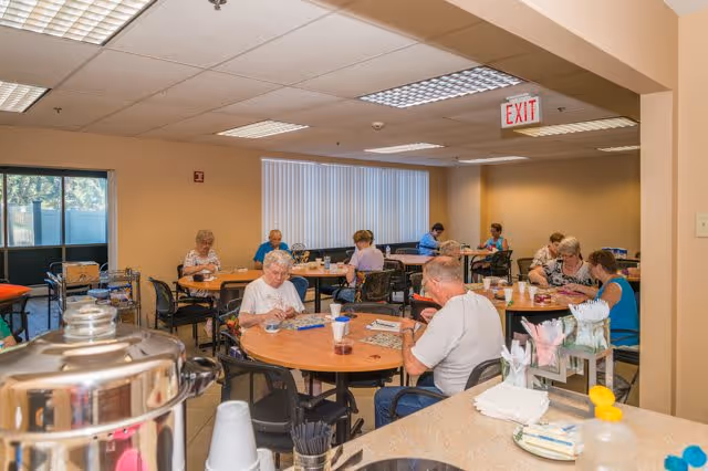 A group of elderly people sitting at round tables in a well-lit room playing games and socializing. The room has beige walls, fluorescent ceiling lights, and large windows with vertical blinds. In the foreground, there is a counter with cups, napkins, and a coffee urn.
