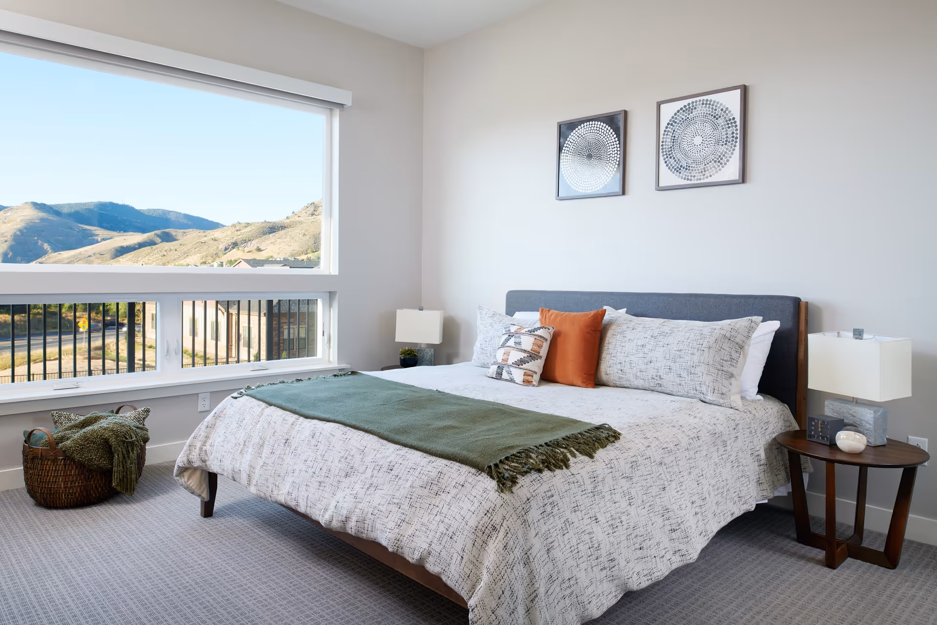 A bright bedroom with a large window showing a scenic mountain view. The bed has a gray upholstered headboard, white and patterned bedding, and three pillows including one orange. There is a green throw blanket at the foot of the bed. A round wooden side table with a modern lamp and decorative items is beside the bed. A woven basket with a green blanket is on the floor near the window. Two framed abstract circular artworks hang on the wall above the bed.