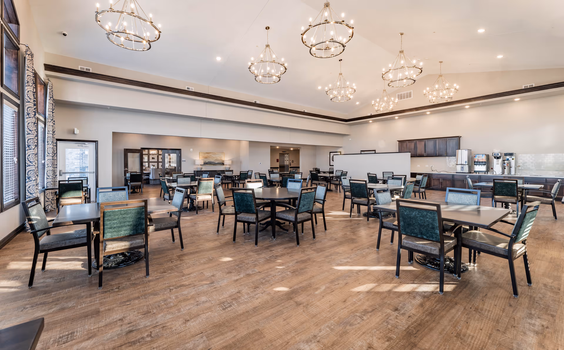 A spacious dining room in Cedarhurst Senior Living of McHenry with multiple tables and chairs arranged neatly on a wooden floor. The room features large windows with patterned curtains, modern chandeliers hanging from the ceiling, and a beverage station with coffee machines and dispensers along one wall.