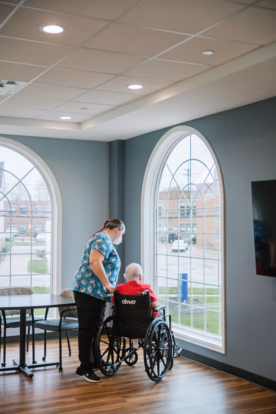 A caregiver wearing a mask stands beside an elderly person in a wheelchair looking out of large arched windows in a bright room with wooden floors and gray walls.
