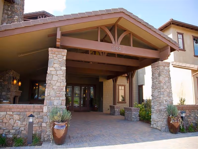 Entrance of a senior living facility with a covered driveway supported by stone pillars, potted plants on either side, and double glass doors leading inside.