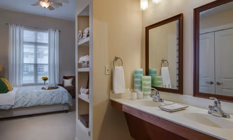 A bright senior living facility room featuring a bed with a blue patterned bedspread and a wooden chair near a window with white curtains. Adjacent to the bedroom is a bathroom area with a double sink vanity, two mirrors, and decorative green and white vases. Shelves with neatly folded towels are visible between the bedroom and bathroom.