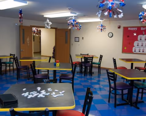 Communal dining/activity room with multiple tables and chairs, festive ceiling decorations, and a birthday bulletin board.
