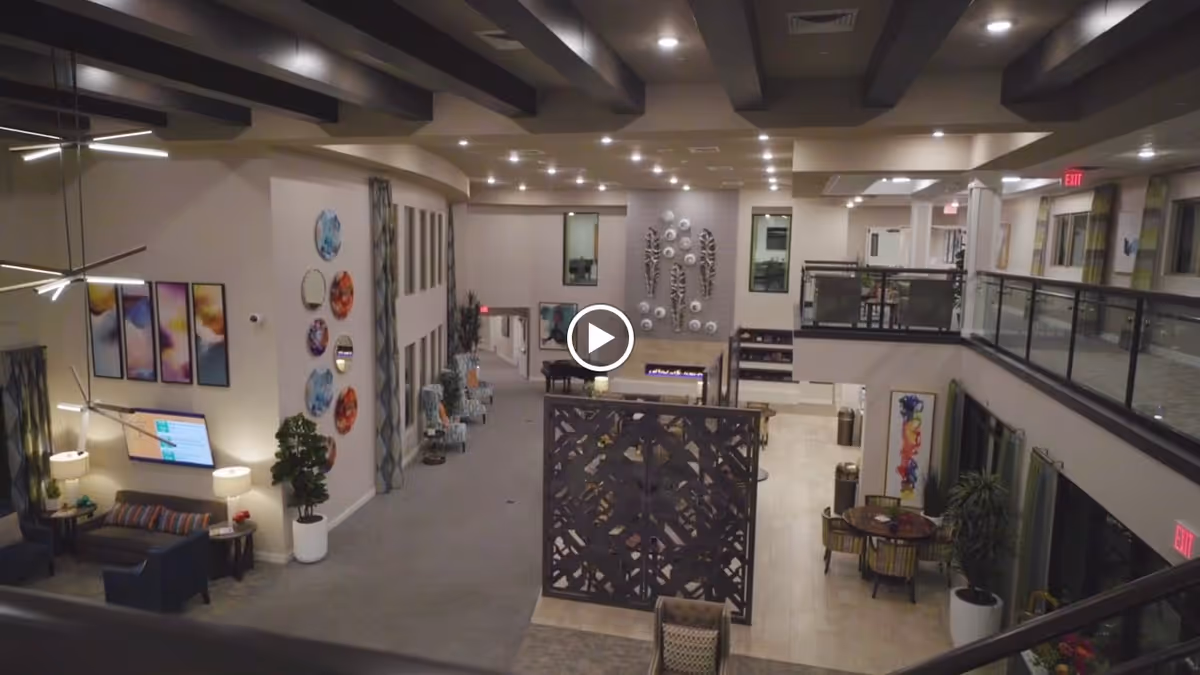 Interior view of a spacious senior living facility lobby with modern decor, including seating areas with chairs and sofas, decorative wall art, plants, and a fireplace. The area has high ceilings with recessed lighting and a mezzanine level with glass railings.