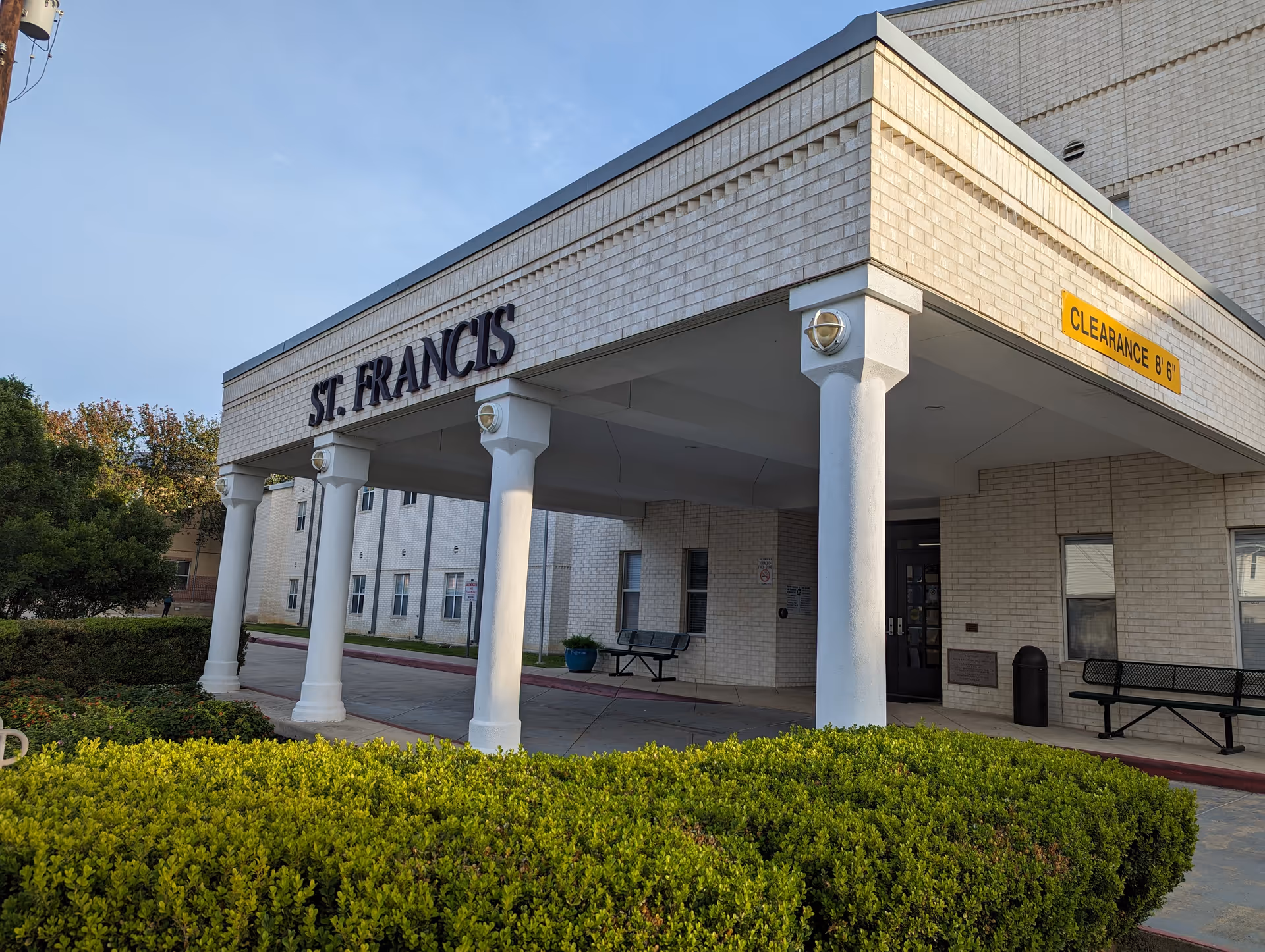 Exterior view of the entrance to St. Francis Nursing Home, featuring a covered drop-off area supported by white columns, with bushes in the foreground and a clearance sign indicating 8 feet 6 inches.