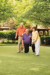 Three elderly people playing golf on a putting green surrounded by trees and greenery. One man in a purple shirt is kneeling and holding a golf ball near the hole, while a woman in a yellow sweater and a man in an orange shirt stand nearby with golf clubs.