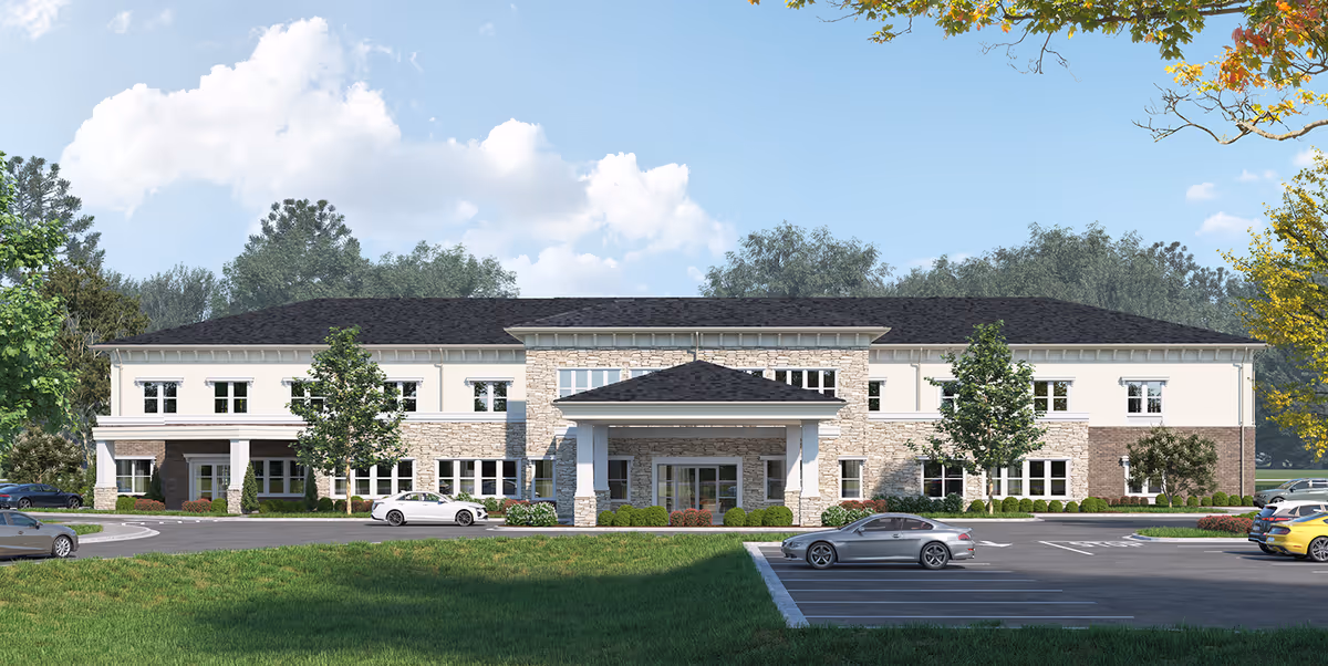Front exterior view of a two-story senior living facility building with a stone and cream facade, a covered entrance, several windows, surrounded by trees and a parking lot with cars under a blue sky with clouds.