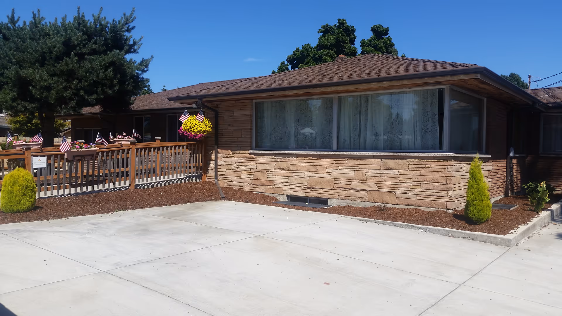 Exterior view of a single-story brick building with a large window covered by lace curtains. There is a wooden fence decorated with small American flags and hanging flower baskets. The area in front of the building is paved with concrete, and there are small shrubs and mulch beds along the building's foundation. The sky is clear and blue.