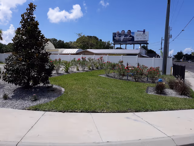 A landscaped outdoor area with a small tree, green grass, and flower beds bordered by gravel. A white fence and a building are visible in the background, along with a billboard and a clear blue sky with some clouds.