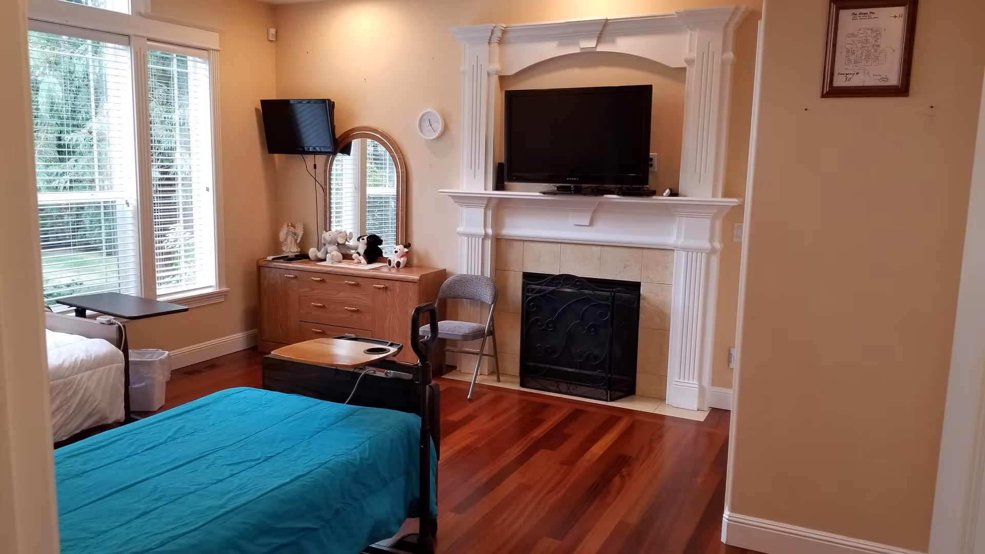 A bedroom with two beds, one with a teal blanket and the other with a white blanket. There is a wooden dresser with stuffed animals on top, a wall-mounted TV above a fireplace with a decorative screen, a chair, and large windows with blinds letting in natural light.