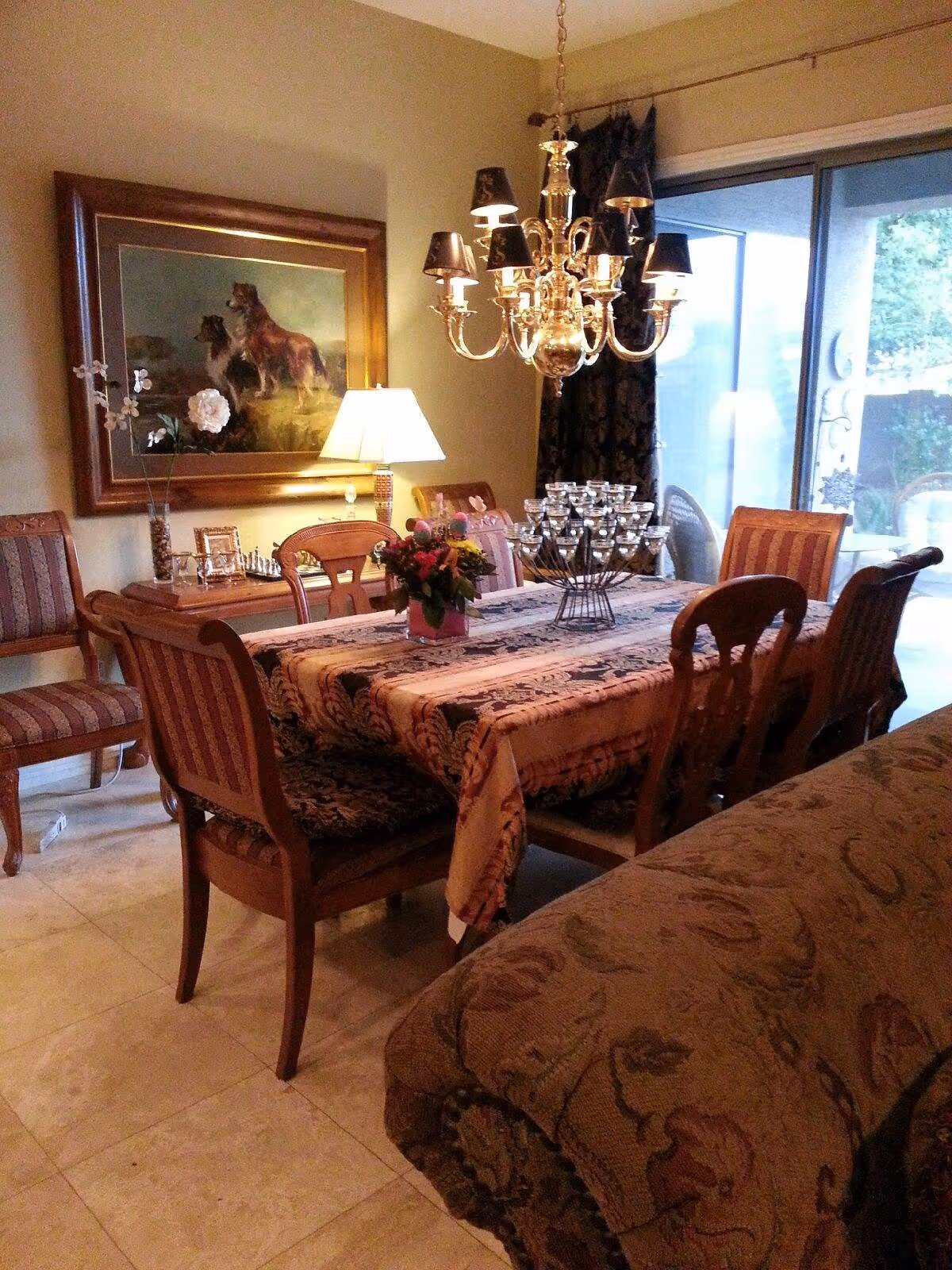 Cozy dining room with a chandelier over a table covered with a patterned tablecloth, wooden chairs, a lamp and framed painting on the wall, and sliding glass doors to a patio.