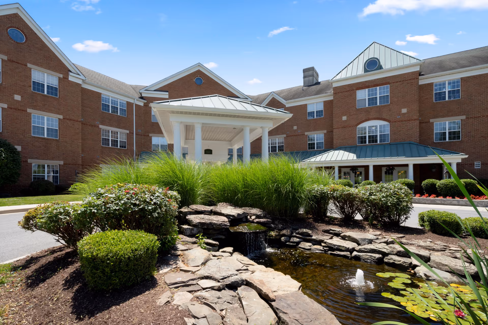 Exterior view of a multi-story brick building with multiple windows and green metal roofs. In front of the building is a landscaped area with bushes, tall grass, rocks, and a small pond with a water fountain. The sky is clear with a few clouds.