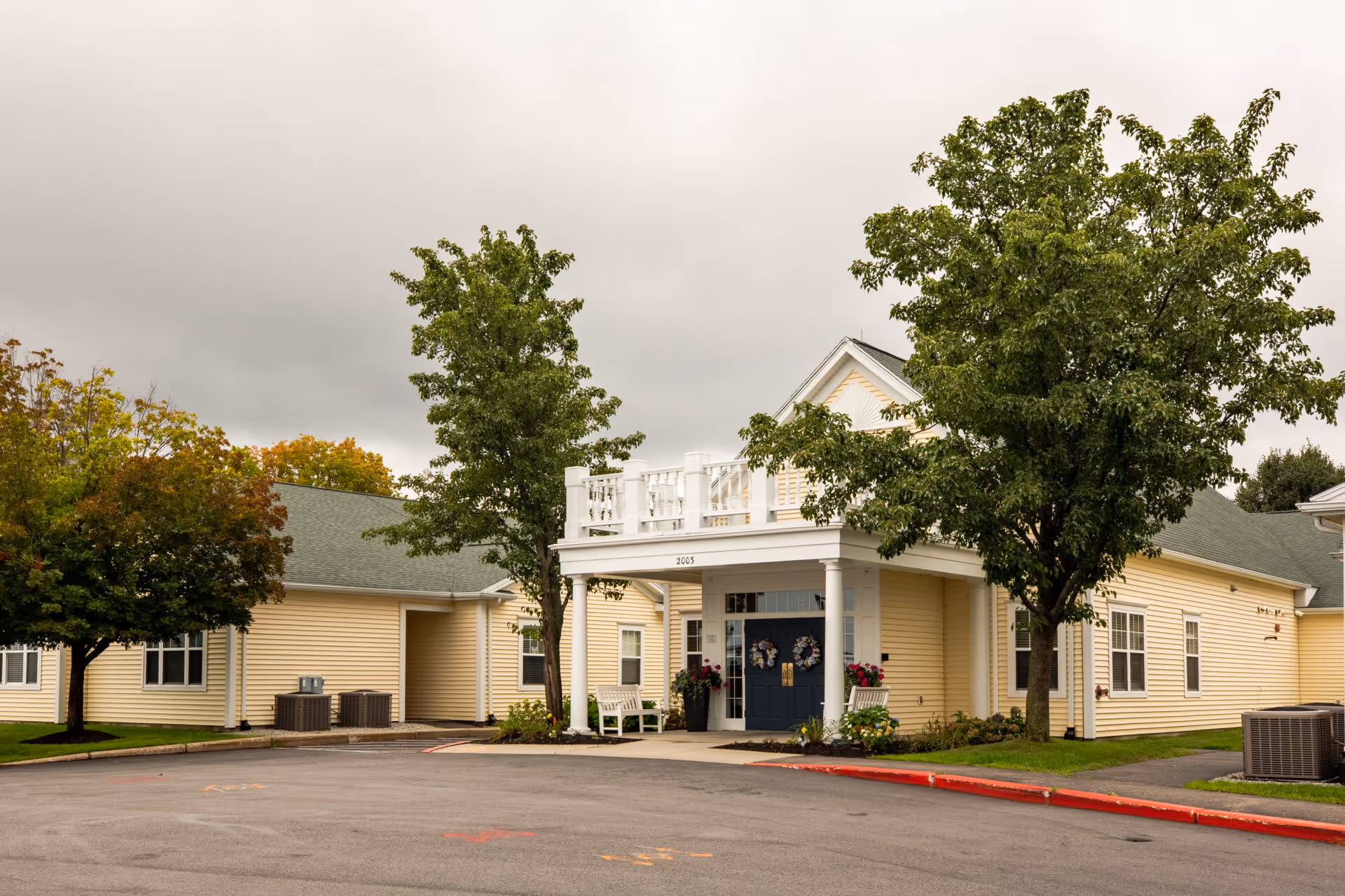 Exterior view of a single-story yellow building with a green roof, white columns, and a blue double door entrance. There are trees and some landscaping around the building, and the sky is overcast.