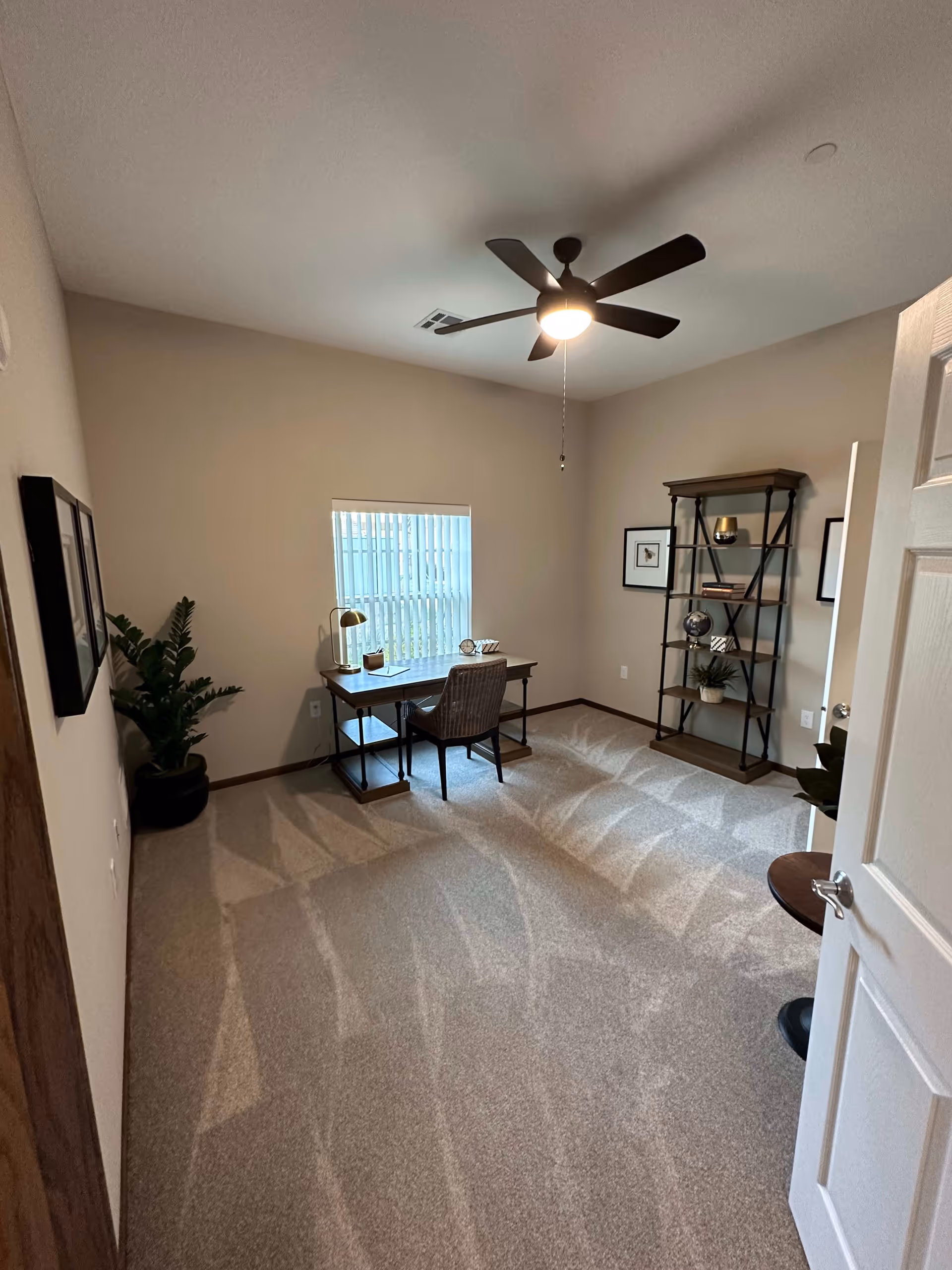 A small, neatly arranged room with beige walls and carpeted floor. The room features a wooden desk with a chair in front of a window with vertical blinds. On the desk are a lamp, a clock, and some decorative items. To the right is a tall wooden shelving unit with various decorative objects and framed pictures on the walls. A ceiling fan with a light is on the ceiling, and a potted plant is in the corner near the desk.