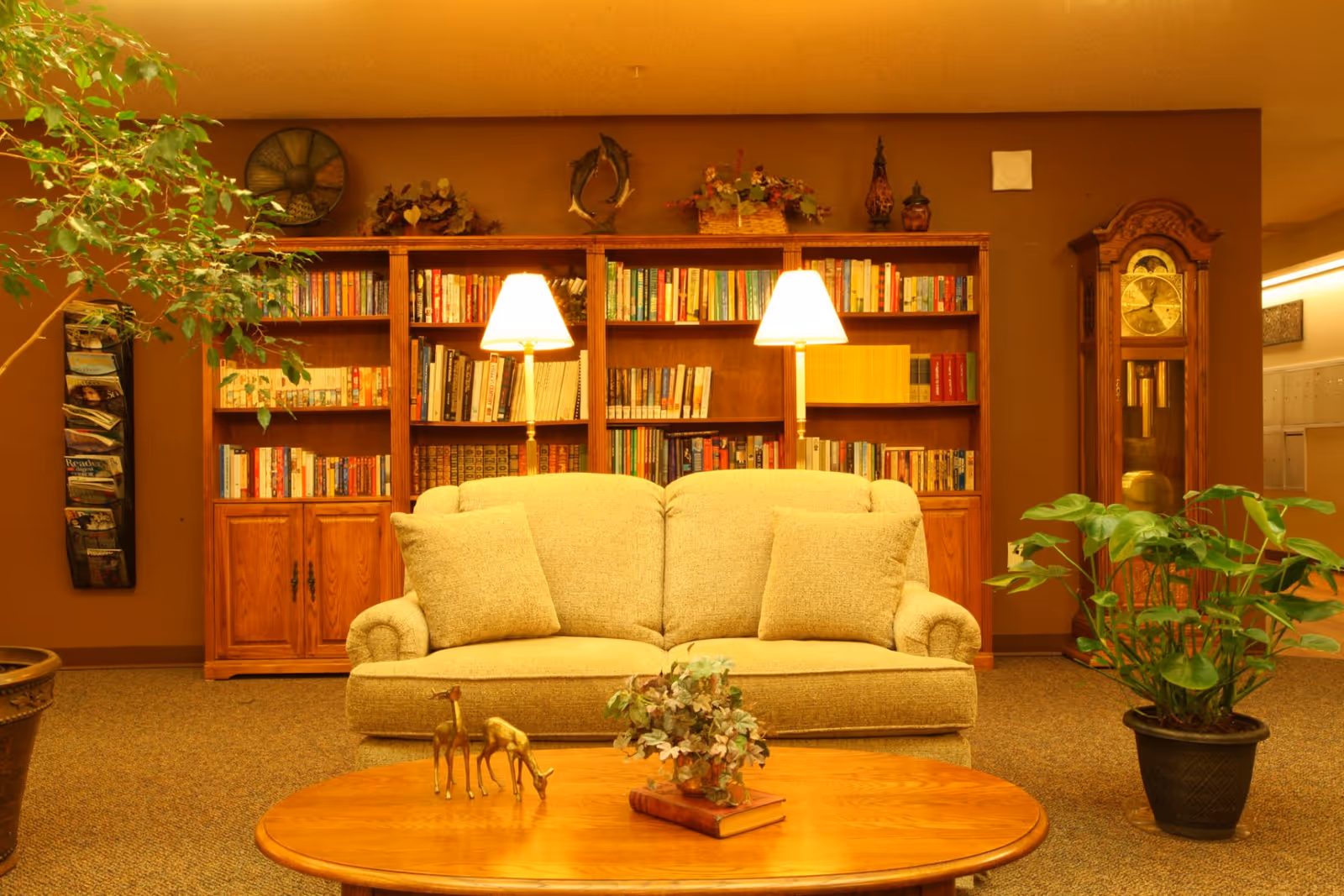Cozy seating area with a beige sofa in front of bookshelves, lamps, a coffee table, plants, and a grandfather clock.
