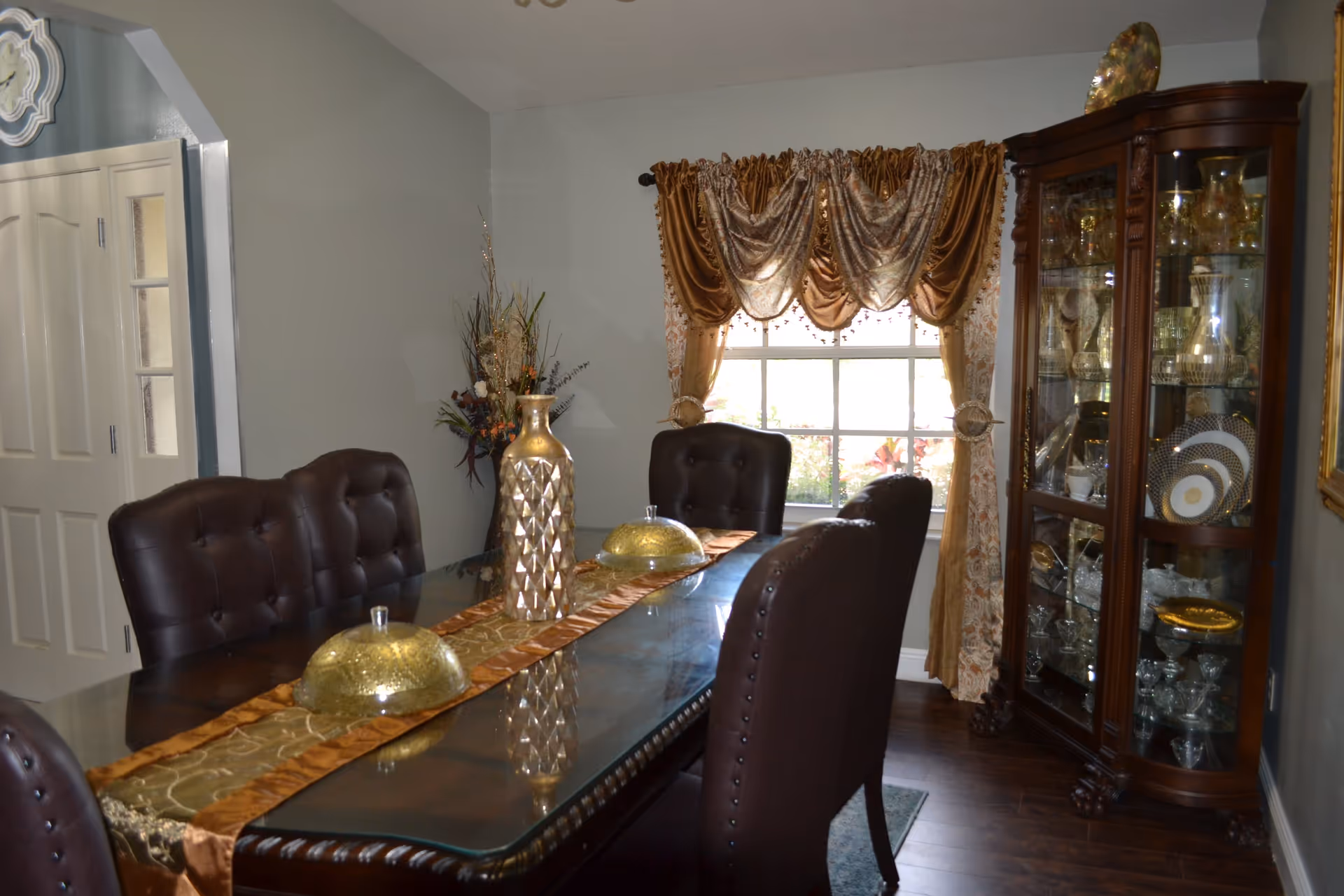 Dining room with a glass-topped table and leather chairs, a decorative centerpiece and gold drapes, and a china cabinet by the window.