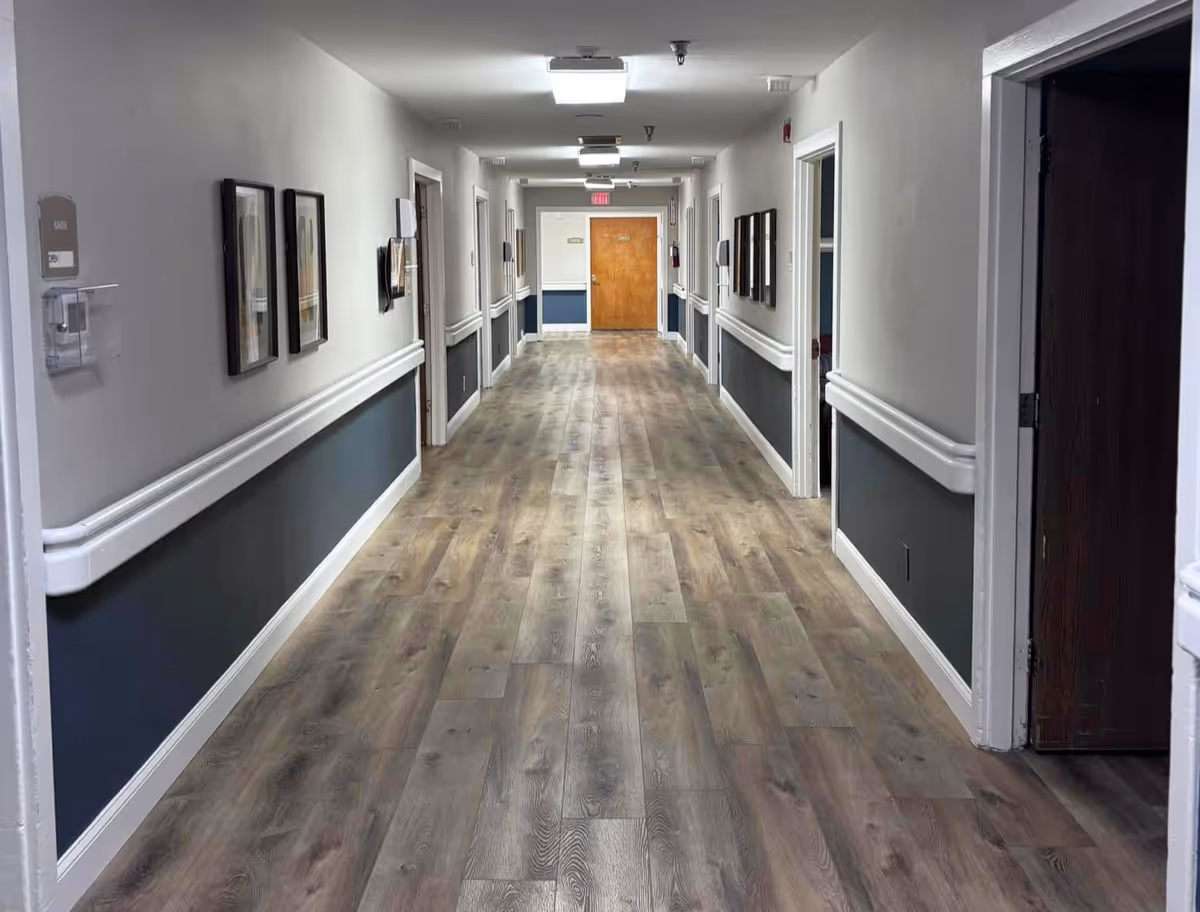 A long, clean hallway in a healthcare facility with wood-patterned flooring, white and dark gray walls, handrails on both sides, framed pictures on the walls, and several doorways leading to rooms. The hallway is well-lit with ceiling lights and has an exit door at the far end.
