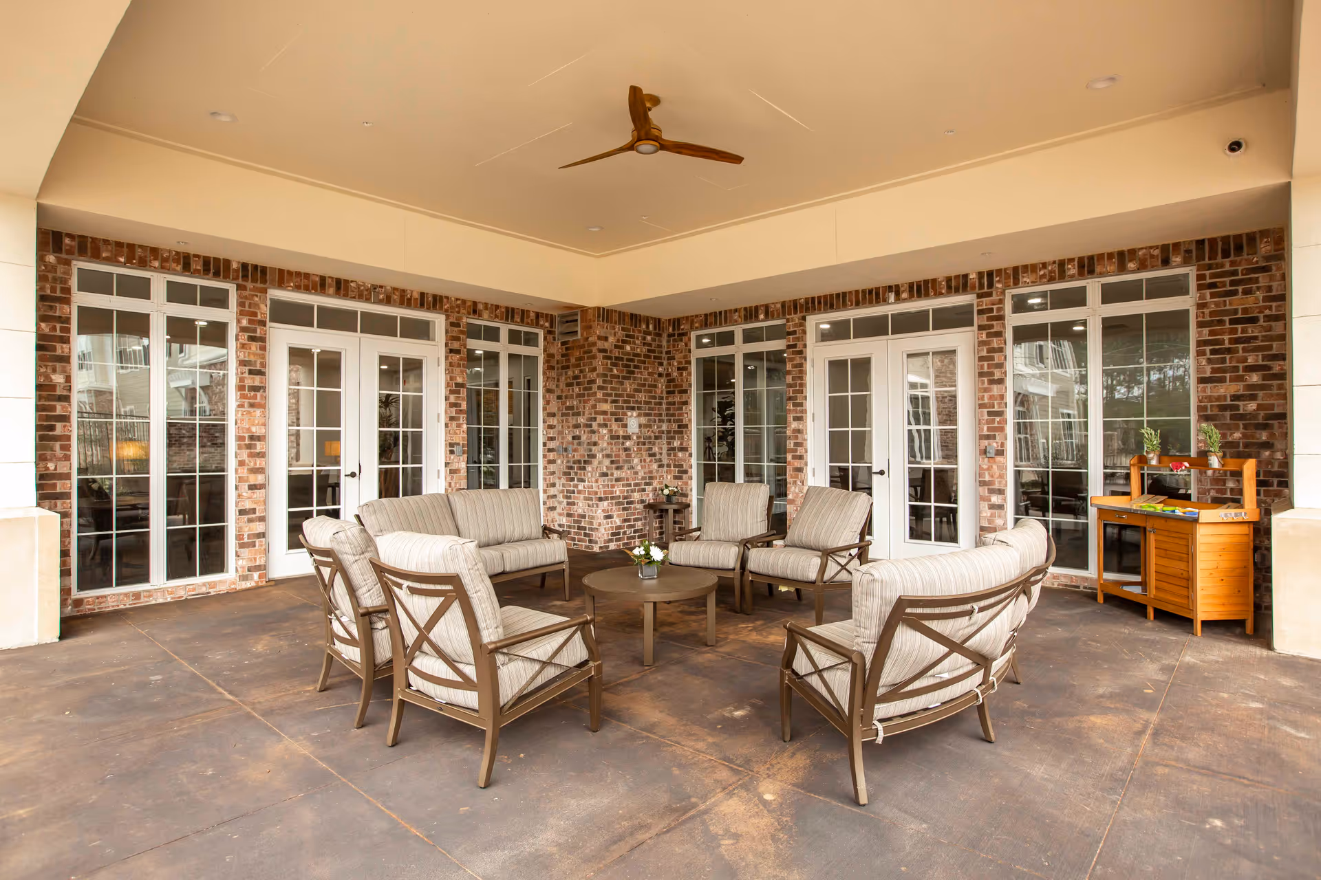 Covered outdoor seating area with cushioned chairs and a round table in front of a brick wall and French doors.