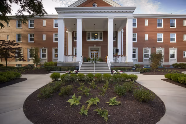 Front exterior view of a senior living facility with a large white columned entrance, brick facade, landscaped garden with shrubs and plants, and a paved walkway leading to the entrance.