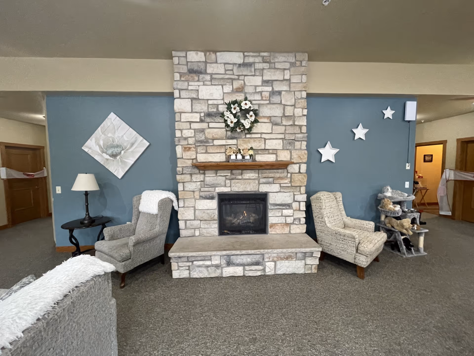 A cozy living room area with a stone fireplace in the center. Above the fireplace is a wooden mantel decorated with a floral wreath and small decorative items. On either side of the fireplace are two armchairs, one gray with a white throw and the other beige with script writing. The wall behind the fireplace is painted blue and decorated with a large flower artwork on the left and three white star decorations on the right. There is a small black side table with a lamp next to the left armchair and a multi-level cat tree with stuffed animals on the right. The room has carpeted flooring and wooden door frames leading to other areas.
