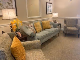 A cozy living room area with a light blue sofa adorned with decorative pillows, two patterned armchairs with yellow cushions, a side table with a framed photo and a lamp, and another lamp on a small table next to the sofa. The walls are decorated with a mirror and framed artwork.