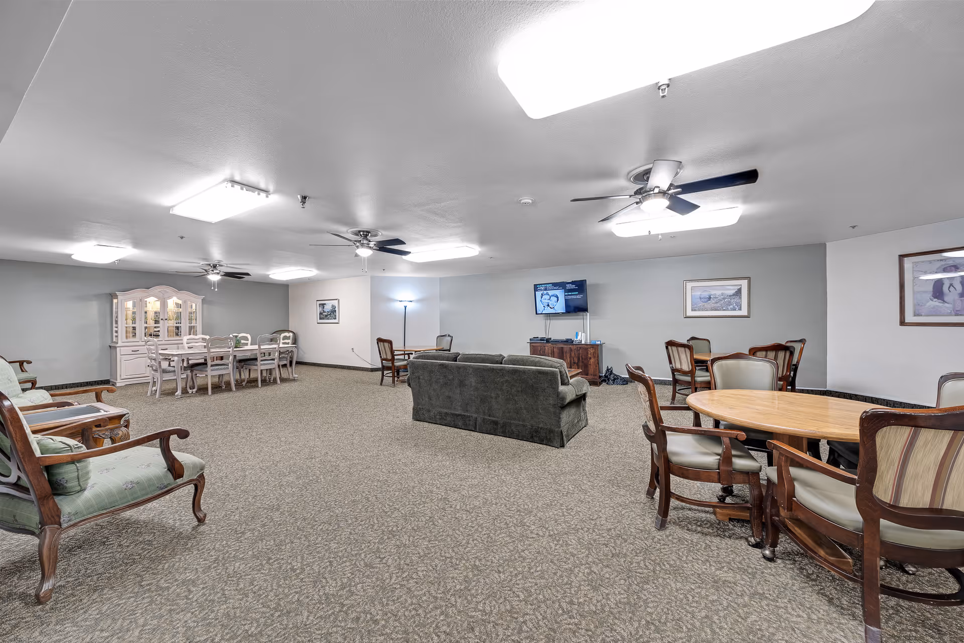 Spacious communal lounge with sofas, multiple dining tables and chairs, a TV on a cabinet, and a china cabinet along the wall.