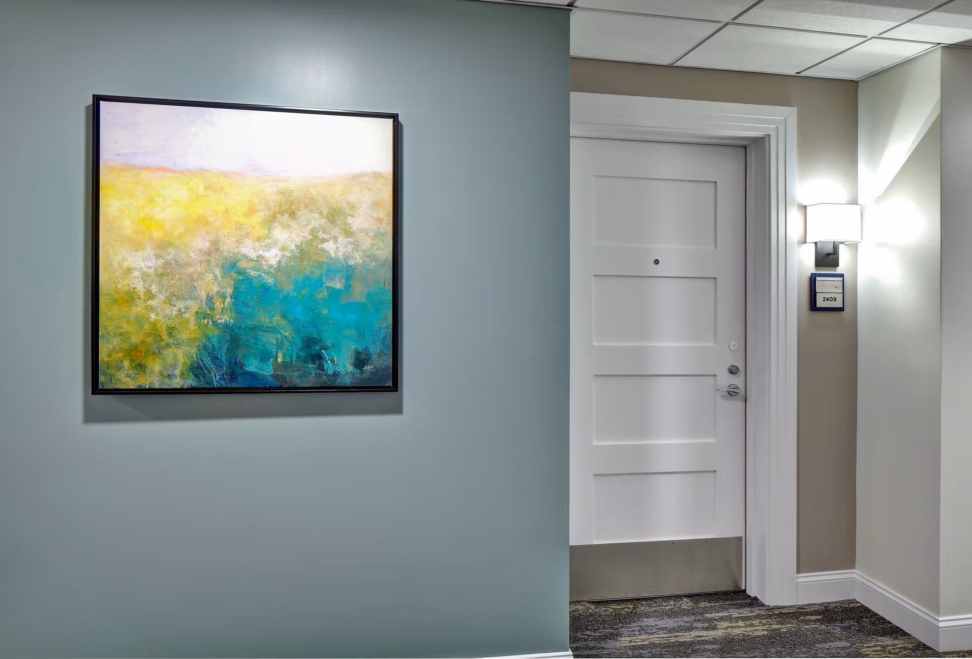Interior hallway of a senior living facility with a white door labeled 2409, a wall-mounted light fixture, and a large abstract painting featuring yellow, green, and blue colors on a light blue wall.