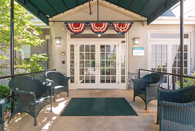 Covered outdoor seating area with four dark wicker chairs arranged around a green mat in front of double glass doors decorated with red, white, and blue bunting. The entrance is shaded by a green awning and surrounded by greenery.