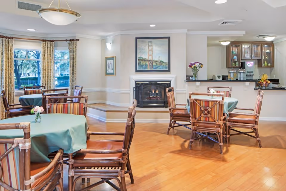 A cozy dining room with wooden floors and several round tables covered with green tablecloths. Each table is surrounded by wooden chairs with striped cushions. There is a fireplace with a painting of the Golden Gate Bridge above it. Large windows with patterned curtains let in natural light. In the background, there is a kitchen area with cabinets, a counter, and some appliances, along with a vase of flowers on the counter.