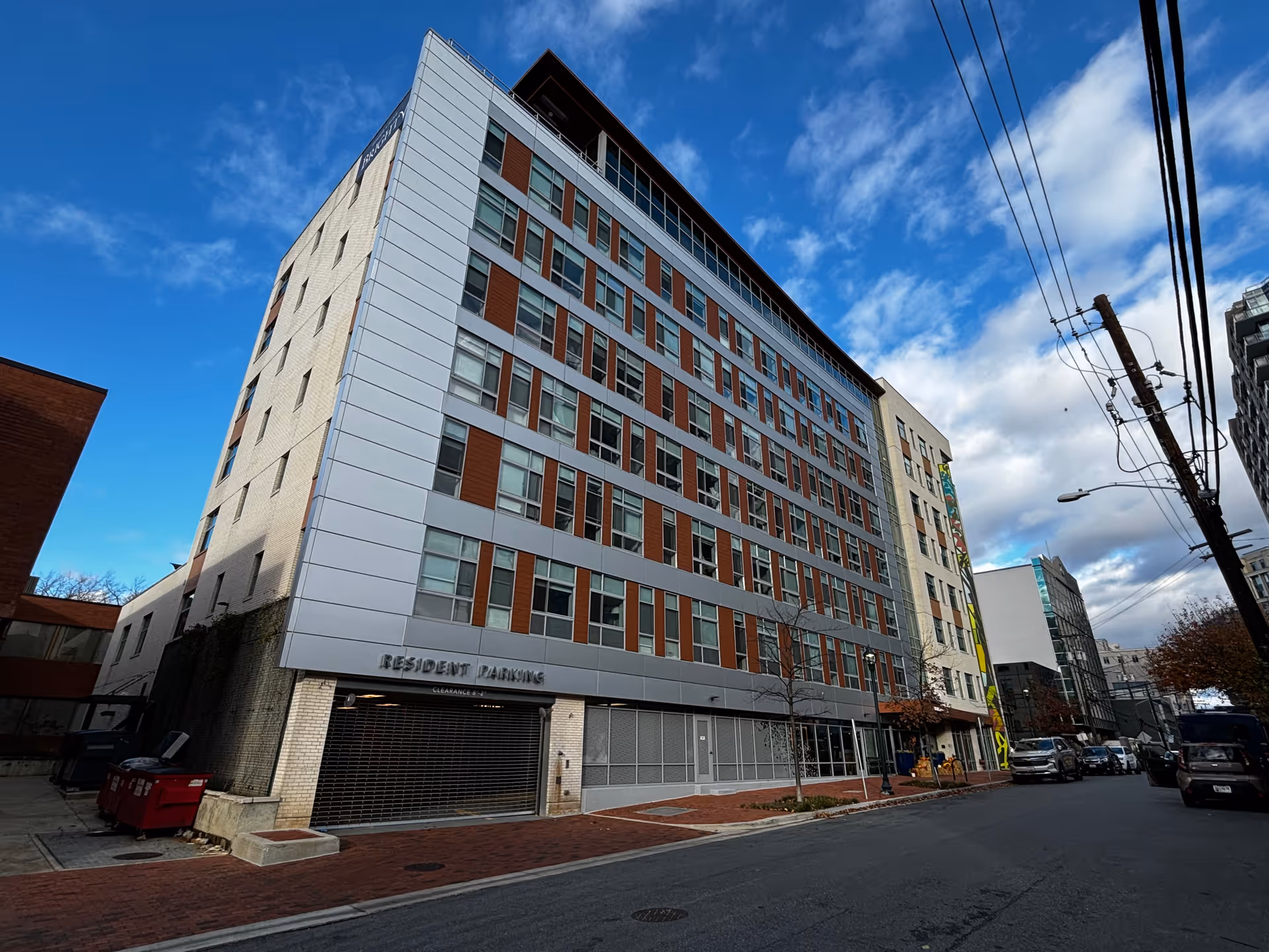Exterior view of a multi-story residential building under a partly cloudy blue sky. The building has a modern design with large windows and a sign indicating resident parking at the ground level. Several cars are parked along the street beside the building.