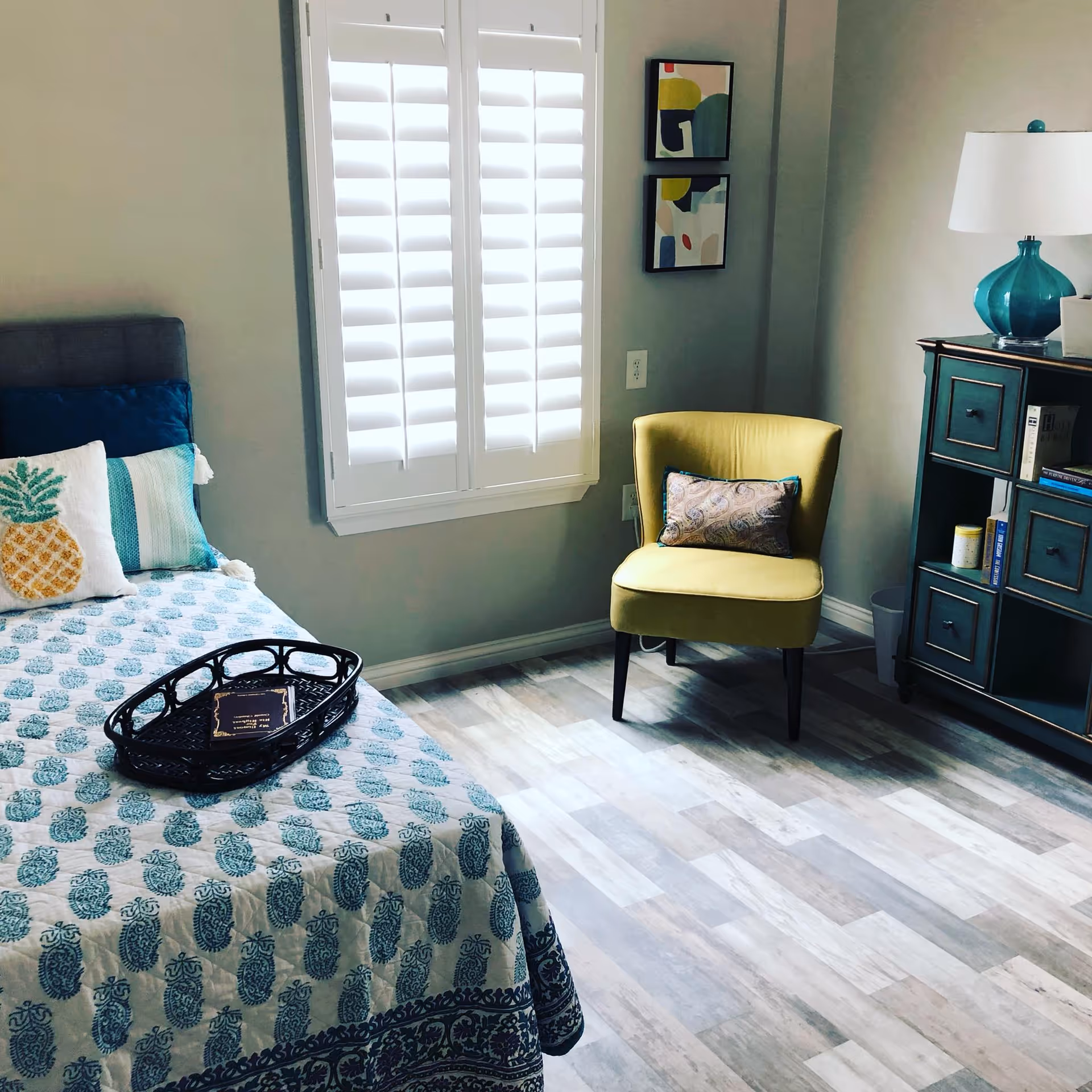 A cozy bedroom with a bed covered in a blue and white patterned quilt and decorative pillows, including one with a pineapple design. A black tray with a book rests on the bed. Next to the bed is a window with white plantation shutters. Across from the bed is a yellow armchair with a decorative pillow. To the right is a teal cabinet with drawers and shelves, topped with a teal lamp and some books. Two abstract art pieces hang on the wall above the chair.